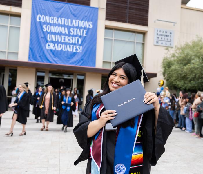 Student in graduation regalia and grad cap holding SSU diploma frame